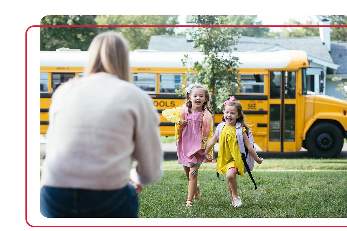 Mother greeting two young daughters as they run from school bus smiling